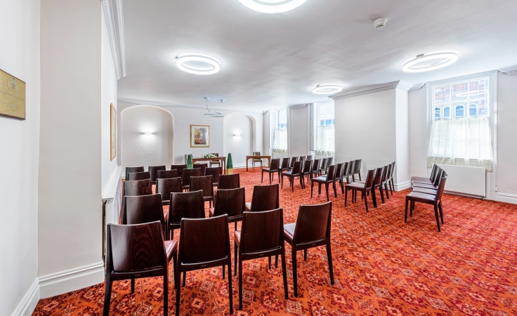 Rows of chairs in front of a table at the end of the room. The table has ornamental plants on either side of it.